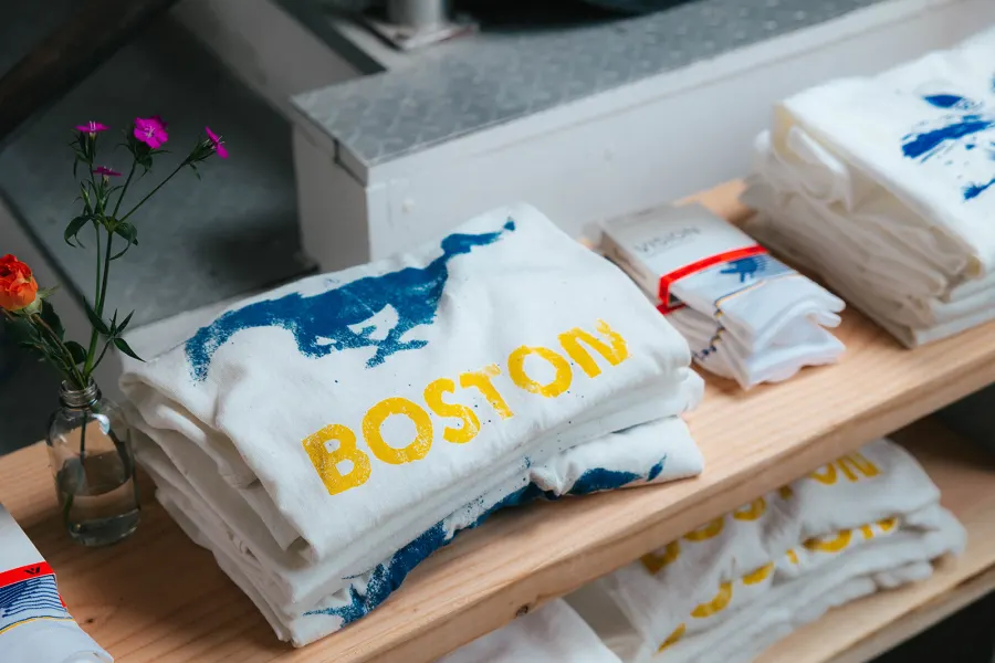 Stacks of freshly screenprinted BOSTON tees folded on a pine shelf next to packaged socks